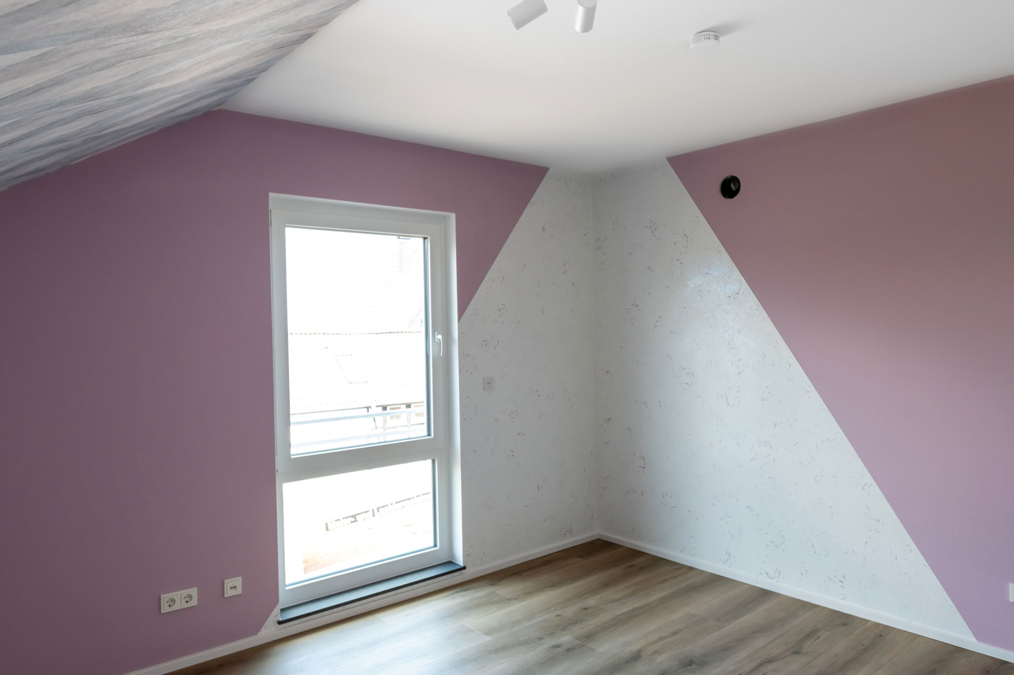 Ein Zimmer im Dachgeschoss in Kreativtechnik mit altrosa Wand auf der in einer Ecke am Fenster zwei weiße Dreiecke zusammen laufen.