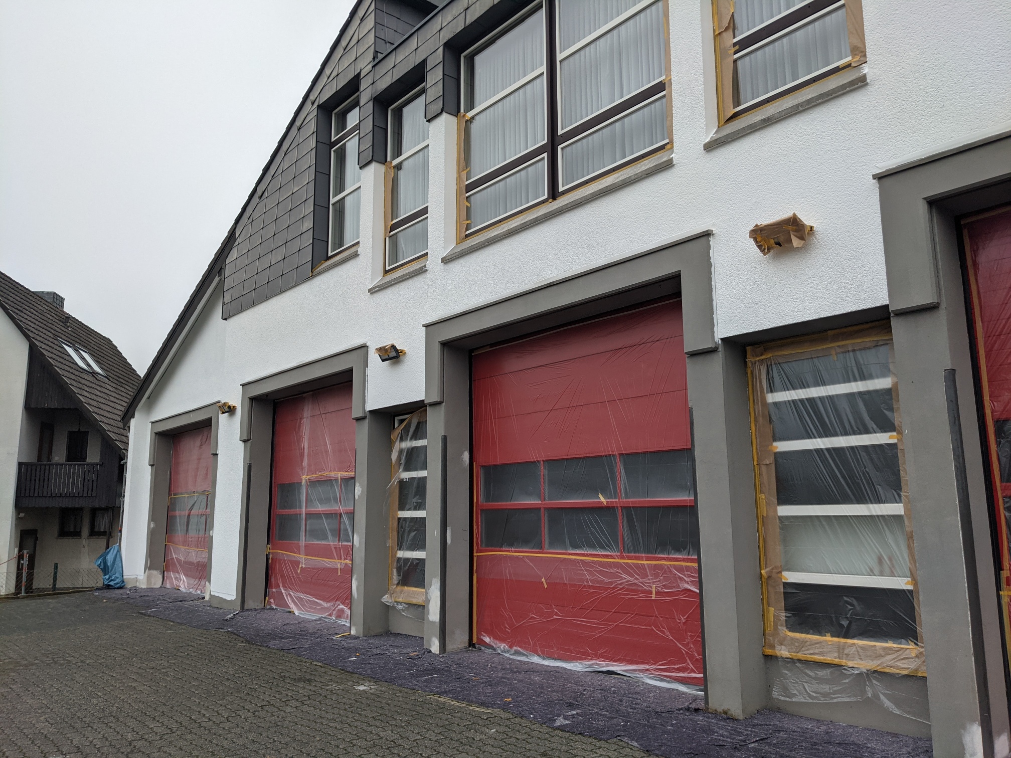 Die für einen Neuanstrich vorbereitete Fassade der Feuerwehr Hennef. Die roten Rolltore und Fenster sind mit Malerkrepp und Folie abgeklebt.