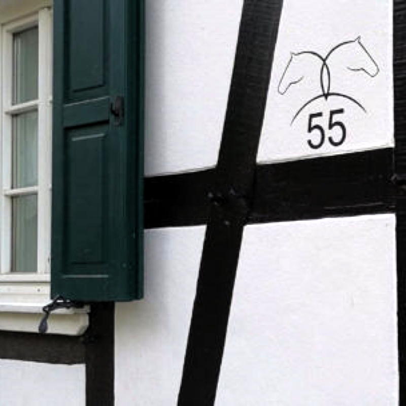 Detail einer sanierten Fachwerkfassade. Grüne Fensterläden und auf ein weißes Gefach wurde die Hausnummer 55 geschrieben, über der sich zwei stilisierte Pferdeköpfe befinden.